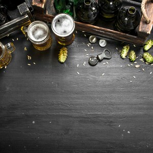 May include: A flat lay of beer bottles, glasses of beer, and hops on a black wooden surface. The beer bottles are in a wooden crate and the glasses of beer are on the surface. There is a bottle opener and bottle caps on the surface.
