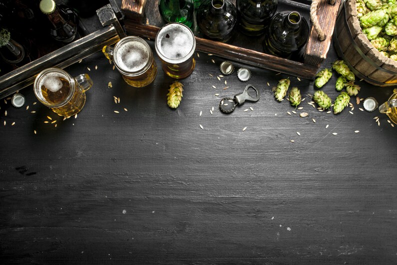 May include: A flat lay of beer bottles, glasses of beer, and hops on a black wooden surface. The beer bottles are in a wooden crate and the glasses of beer are on the surface. There is a bottle opener and bottle caps on the surface.