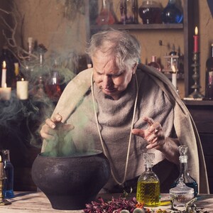 May include: An older man in a brown robe stirs a cauldron with a green liquid and smoke rising from it. He is surrounded by candles, bottles, and other potion-making supplies.