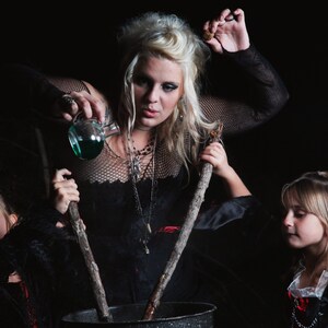 May include: A woman dressed as a witch pours a green liquid into a cauldron while two children dressed as witches stir the cauldron with wooden sticks.