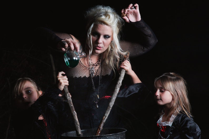 May include: A woman dressed as a witch pours a green liquid into a cauldron while two children dressed as witches stir the cauldron with wooden sticks.
