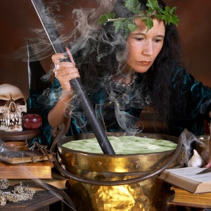 May include: A woman wearing a green velvet robe and a wreath of leaves stirs a cauldron with a wooden spoon. The cauldron is filled with a green liquid and steam is rising from it. There is a skull, a book, and other potion bottles on the table.