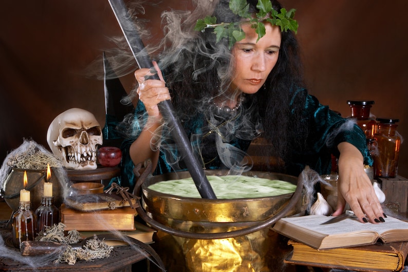 May include: A woman wearing a green velvet robe and a wreath of leaves stirs a cauldron with a wooden spoon. The cauldron is filled with a green liquid and steam is rising from it. There is a skull, a book, and other potion bottles on the table.