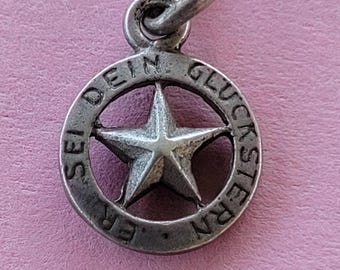 May include: A small, silver-toned pendant featuring a five-pointed star within a circular frame. The frame has the words "SEI DEIN GLÜCKSTERN" engraved around the star. The pendant has a small loop at the top for attaching to a chain or bracelet.
