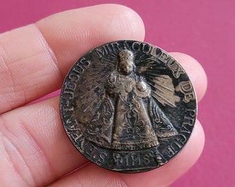 May include: A round, bronze-colored medal with a raised image of a person in a robe and a crown. The medal has the text "JESUS MIRACULOS DE CHIPA" around the edge.
