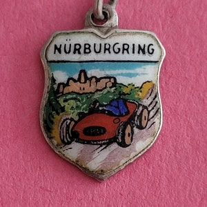 May include: Silver charm with a white enamel shield featuring a red race car driving on a road with a green hill and a castle in the background. The text "NÜRBURGRING" is written in black on the shield.