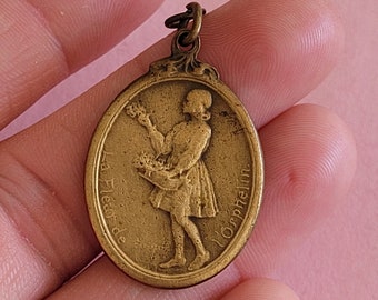 May include: A gold-coloured metal pendant featuring a raised image of a girl holding flowers. The pendant is oval-shaped and inscribed with the text "La Fleur de l'Orphelin" around the edge.