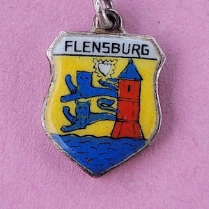 Puede incluir: Un amuleto de plata con un escudo de esmalte azul y amarillo que representa el escudo de armas de Flensburg, Alemania. El escudo muestra una torre roja con tres gatos azules trepando por ella.