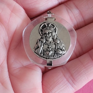 Op de afbeelding: Zilverkleurige religieuze hanger met een afbeelding van een figuur met een halo, geplaatst in een heldere, afgeronde plastic behuizing. De hanger heeft een kleine lus aan de bovenkant om op te hangen. De achtergrond is effen roze.