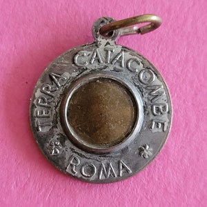 Op de afbeelding: Ronde zilverkleurige hanger met de woorden "TERRA CATACOMBE ROMA" gegraveerd rond de rand. De hanger heeft een klein lusje aan de bovenkant om een ketting aan te bevestigen.