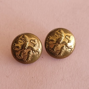 Könnte beinhalten: Zwei goldfarbene Knöpfe mit einem erhabenen Design eines Löwen. Der Löwe blickt nach rechts und trägt eine Krone auf dem Kopf.