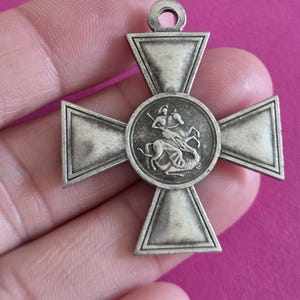 May include: A silver-toned metal cross pendant featuring a central circular medallion with a relief depiction of a figure on horseback slaying a dragon. The cross has four flared arms with raised borders and a small circular loop at the top for hanging.