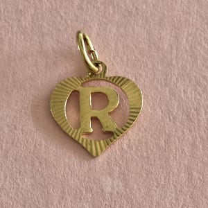 May include: A gold-toned metal charm in the shape of a heart with a cut-out letter R in the centre. The heart features a textured, sunburst-like pattern radiating from the centre letter toward the outer edges, and includes a small jump ring at the top.