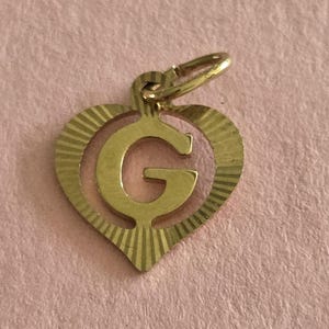 Op de afbeelding: Een goudkleurige metalen bedel in de vorm van een hart met een uitgesneden letter G in het midden. Het hartframe is voorzien van een gestructureerd, in zonneschijnstijl gegraveerd patroon dat vanuit het midden uitstraalt, met een ringetje.