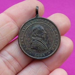 Puede incluir: Una medalla de bronce con el retrato de un hombre que lleva un sombrero y una túnica. La medalla tiene el texto "XII PONTIFICE MAXIMO" y "ROMA" alrededor del borde.