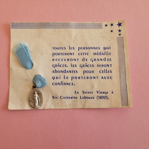May include: A vintage religious medal with a blue ribbon attached to a card with French text. The medal features an image of a saint. The card has text and star illustrations.