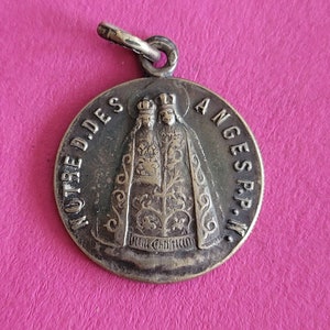 Puede incluir: Un colgante de metal plateado con una imagen religiosa de dos figuras con coronas, con el texto "NOTRE D. DES ANGES P.P.W." alrededor del borde.