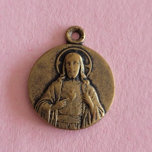 May include: A gold-toned metal pendant featuring a relief image of Jesus Christ with a heart on his chest. The pendant is circular and has a loop at the top for attaching to a chain.