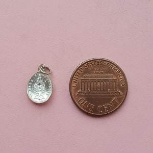 May include: A silver charm with a detailed image of a building, possibly a church, with the words "Ave Maria" inscribed around the edge. The charm is next to a US penny for size comparison.