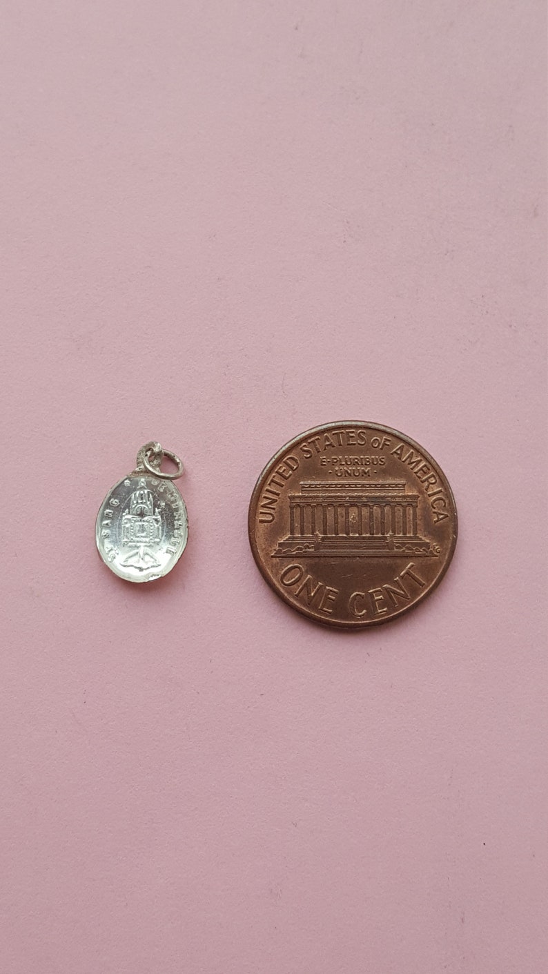 May include: A silver charm with a detailed image of a building, possibly a church, with the words "Ave Maria" inscribed around the edge. The charm is next to a US penny for size comparison.