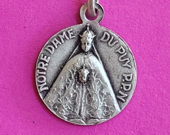 May include: A silver-colored pendant with the text "NOTRE DAME DU PUY P.P.N." encircling a detailed image of a robed figure. The pendant has a loop at the top for a chain, set against a pink background.