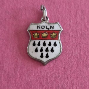 Könnte beinhalten: Ein silberfarbener Metallanhänger in Wappenform mit weißer Emaille und schwarzen Tropfenmotiven, einem roten Querbalken mit drei gelben Kronen sowie dem Schriftzug KÖLN in schwarzen Buchstaben im oberen Bereich mit einer kleinen Metallöse.