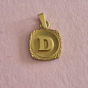May include: A gold-toned pendant featuring a square shape with rounded corners and a textured, cross-hatched border. The centre of the pendant displays a raised, polished capital letter D. A small, integrated bail is attached to the top.