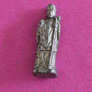 Op de afbeelding: Een klein, zilverkleurig metalen beeldje dat een staand figuur voorstelt met een hoge, gestructureerde hoed en lange, gedrapeerde gewaden. Het figuur houdt een hoge, slanke staf in één hand en staat op een platte, rechthoekige basis.