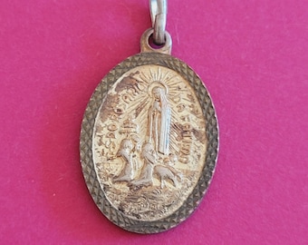 Può includere: Pendente ovale in argento con una rappresentazione della Vergine Maria e due bambini. Il pendente ha un bordo a forma di diamante e le parole "Ave Maria" e "Gratia Plena" sono incise intorno all'immagine.