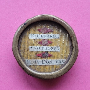 Op de afbeelding: Een klein, rond, antiek religieus relikwie met een goudkleurig metalen frame. De binnenkant heeft een gele achtergrond met drie rechthoekige labels met de namen "B. GERARDI", "S. ALPHONSI" en "R. P. DONDERS". Rode en witte decoratieve elementen zijn ook aanwezig.