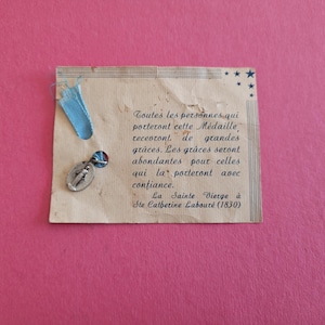 May include: A vintage religious medal with a blue ribbon attached to a worn paper card. The card has French text and small blue stars. The medal is silver-toned and oval-shaped.