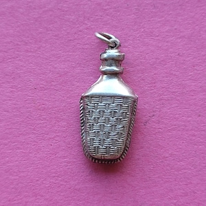 Op de afbeelding: Zilverkleurige hanger in de vorm van een fles, met een geweven mandontwerp. De fles heeft een gedetailleerde dop en een lus om aan een ketting te bevestigen. De hanger is van metaal en staat tegen een roze achtergrond.