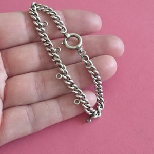 Op de afbeelding: Een zilverkleurige metalen kettingarmband met een reeks kleine, ronde verbindingsringen die met tussenpozen aan de schakels van de stoepketting zijn bevestigd. De armband is beveiligd met een ronde veerringsluiting voor een functionele sluiting.