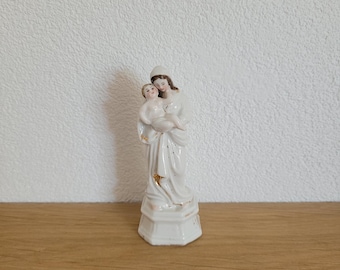 May include: White porcelain figurine of the Virgin Mary holding a baby Jesus. The figurine is detailed with gold accents and has a simple, elegant design.
