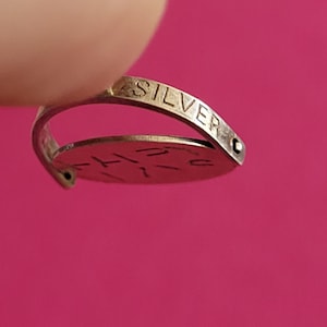 Op de afbeelding: Een zilverkleurige metalen ring met een dunne, gebogen band waarop het woord SILVER op de buitenkant is gegraveerd. Een platte, ronde schijf is aan de band bevestigd en toont gedeeltelijk zichtbare gegraveerde tekst op het oppervlak.