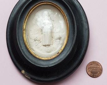 May include: A white cameo of a person in a long robe, framed in a black oval frame with a gold trim.