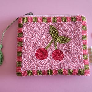 Può includere: Portamonete rosa con perline con un disegno di ciliegie. La borsa ha una forma quadrata con uno sfondo di perline rosa e un bordo di perline rosa e verdi. Un nappa verde pende dalla cerniera. Una moneta è accanto alla borsa.