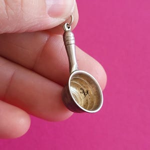 Op de afbeelding: Een kleine zilverkleurige metalen bedel in de vorm van een miniatuur steelpan met een lange steel en een ronde kom. De binnenkant van de kom is voorzien van een reeks concentrische cirkelvormige ribbels en een klein donker vlekje.
