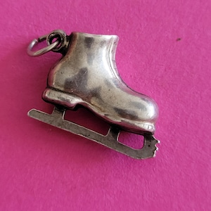 Op de afbeelding: Een zilverkleurige metalen bedel in de vorm van een schaats, met een glad laarsontwerp, een zichtbaar ijzer aan de onderkant en een kleine ronde ring aan de bovenkant om aan een armband of ketting te bevestigen.