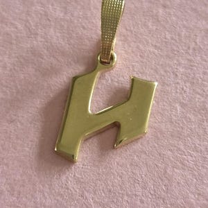 May include: A gold-coloured pendant shaped like the letter H in a gothic-style font. The pendant features a smooth, polished surface and includes a textured, rectangular bail at the top for attaching to a chain or necklace cord.