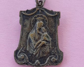 May include: Antique silver-toned pendant featuring a detailed relief of a woman holding a child, framed within an ornate, shield-shaped border. The pendant has a loop at the top for a chain.