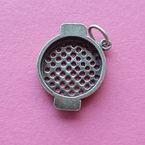 Puede incluir: Un colgante plateado con un diseño circular perforado. El colgante tiene un pequeño lazo en la parte superior para una cadena y dos extensiones rectangulares en lados opuestos. El fondo es de color rosa sólido.