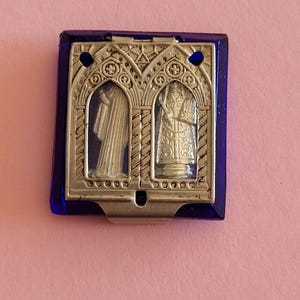 May include: A rectangular, blue-edged religious icon with a silver-colored metal frame. The frame features two arched windows with religious figures inside. The icon has two small holes at the top and a metal clip at the bottom.