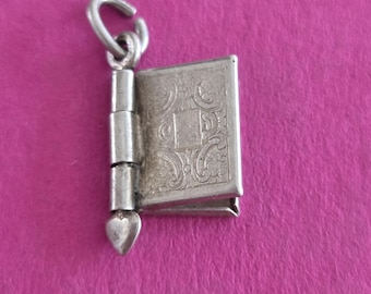 May include: A silver-toned metal charm shaped like a small book with an intricate embossed floral pattern on the cover. The charm features a vertical spine with a small heart-shaped detail at the bottom and a circular jump ring at the top.
