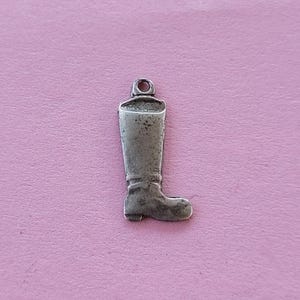 May include: A silver-colored charm in the shape of a riding boot. The boot has a detailed design, including the shape of the foot and the top of the boot. The charm has a small loop at the top for attaching to a necklace or bracelet. The background is a solid pink color.