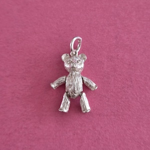 Op de afbeelding: Een zilverkleurige metalen bedel in de vorm van een teddybeer met textuurdetails van vacht, kleine ronde oortjes en een cirkelvormige ring aan de bovenkant van het hoofd om op te hangen. De beer heeft uitgestrekte armen en benen.