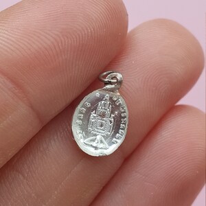 May include: Silver oval charm with a religious image of a church and the text "St. Sang de Christ" around the edge.