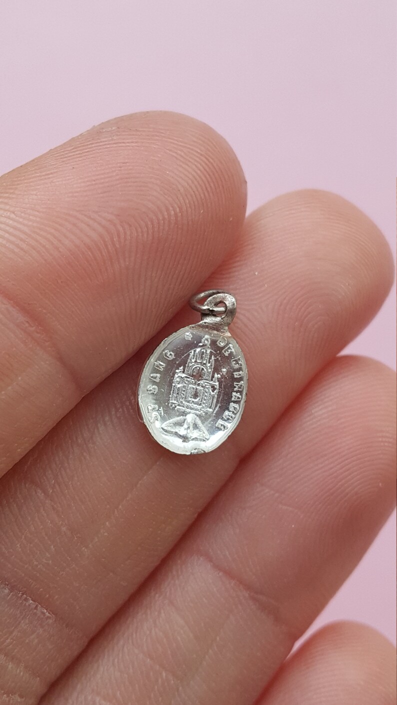 May include: Silver oval charm with a religious image of a church and the text "St. Sang de Christ" around the edge.