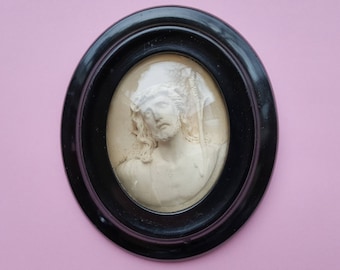 May include: A black oval frame with a white cameo of a man's face. The cameo is raised and has a three-dimensional effect.