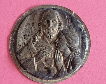 May include: A circular, silver-toned religious medal featuring a detailed relief of a figure holding a chalice and a person with clasped hands. The medal has an aged appearance and is set against a pink background.